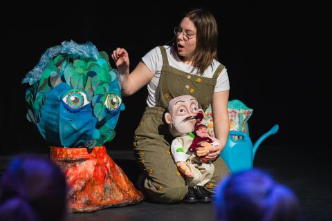 Une grande boule bleu et verte avec des yeux, une marionnette d'un humain en complet cravate et une d'un humain avec une tuque. La manipulatrice au cheveux châtain a une exclamation de surprise sur son visage.  