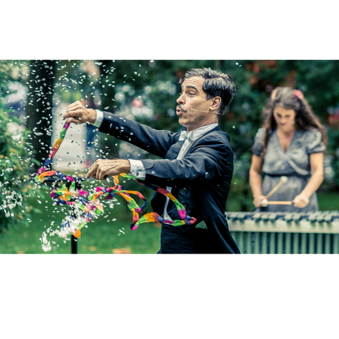 Un monsieur entrain de faire un tour de magie avec des bulles et derrière lui une dame qui joue un instrument de musique