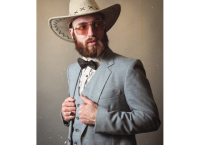 L'artiste avec un chapeau de cowboy sur la tête et portant une veste grise avec un noeud papillon.