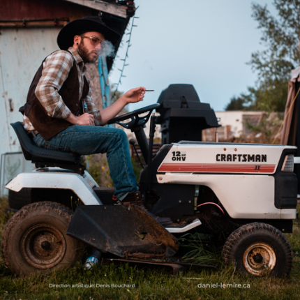 L'artiste Menoncle Jason assis sur un petit tracteur et fumant une cigarette avec un chapeau de Cowboy sur la tête.